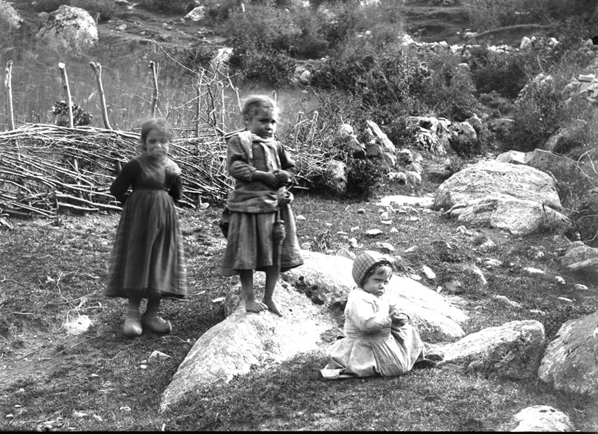 Tres nenetes jugant fora del poble. La mitjana, descalça sobre la roca, fila la llana.