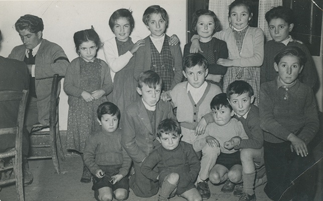 <p>Grup de nens i nenes a la Taverna Casa Negret d’Isavarre. A l’esquerra es veu Pepe de Ramón jugant a la brisca. D’esquerra a dreta fila superior: Pepe de Ramón, Pepita de Ricart, noia de Berrós, Emília ”Milieta” de Casa Miqueu, Neus de Casa Carles, Inés de casa Negret i Llorenç Costança. Fila central, esquerra a dreta: Sintet de casa Piarró, Josep de casa Carles, Manolin de casa Miqueu, ? germà Manolin, Enriquet de casa Miqueu Josep Constança i Josep Mº de casa Negret (nen en braços)</p>